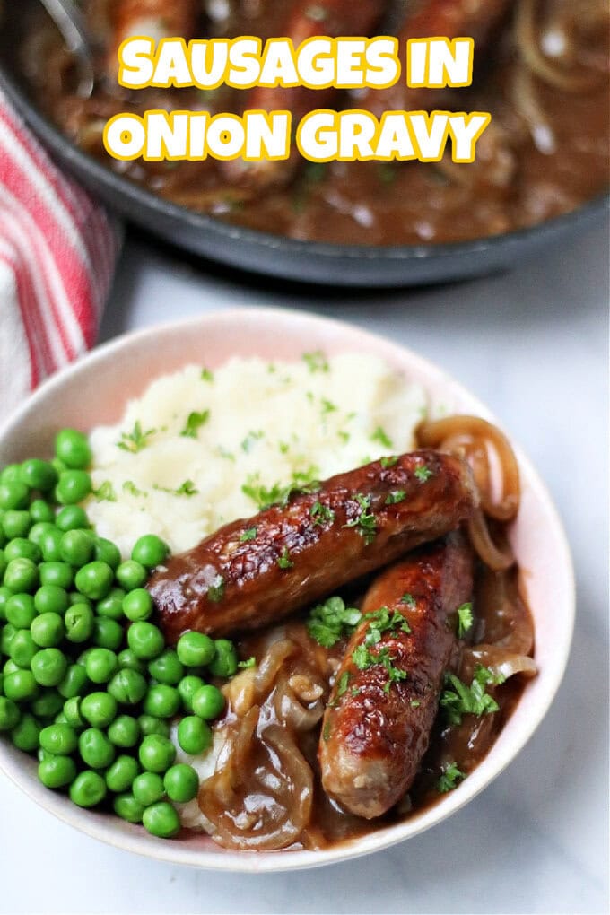 Sausages In Onion Gravy in a light pink bowl with mashed potato, a side serving of green peas and garnished with freshly chopped parsely.