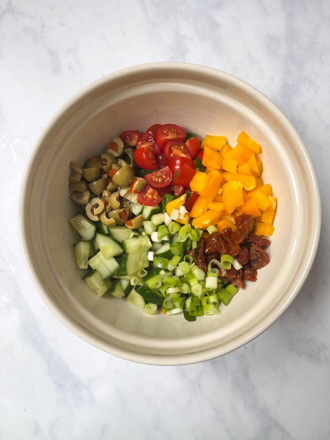 lettuce, cherry tomatoes, cucumber, yellow pepper, olives, sun dried tomatoes and spring onion chopped in a white ceramic bowl