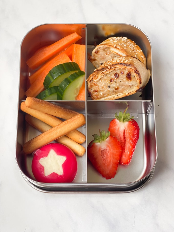 chicken sausage rolls in a compartment lunch box with fresh veggies, fruit, mini breadsticks and a portion of cheese
