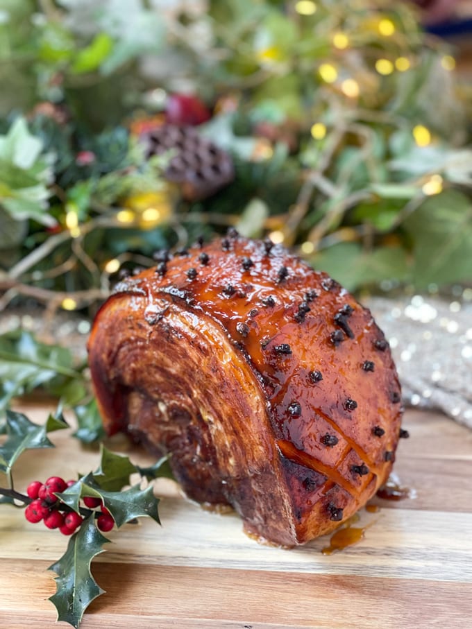 Christmas Ham In Coke with a sticky glaze, served on a wooden board with festive decorations in the background.