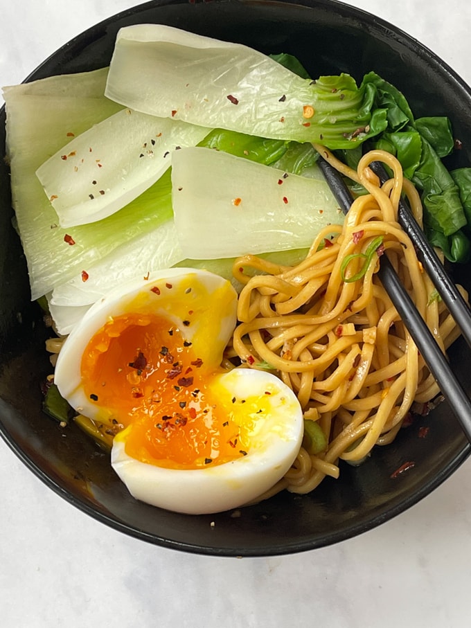 Hot oil noodles with pak choi and an egg served in a black bowl with chopsticks