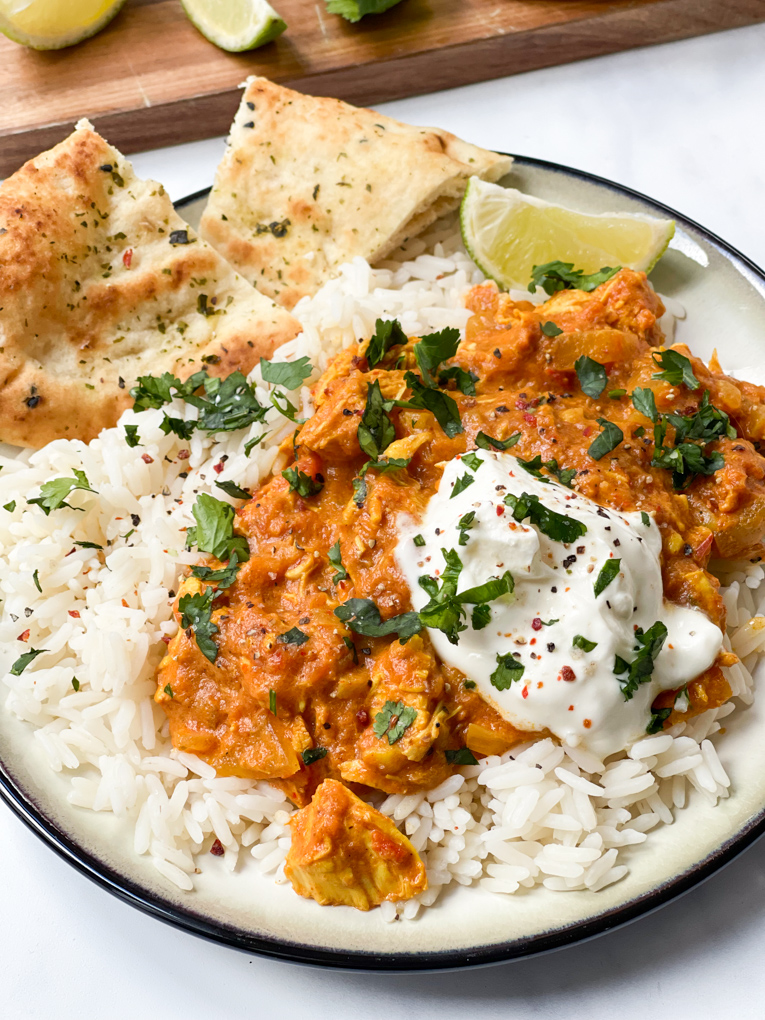 Slow cooker chicken Tikka Masala served on a large plate with white rice and naan bread pieces. Garnished with a slice of lime and a sprinkle of coriander.