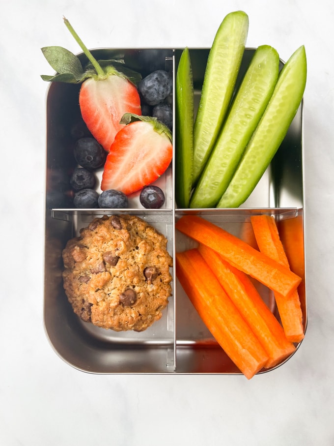 chunky chocolate chip oat cookies in a metal lunch box with strawberries, blueberries, sliced cucumber and carrot batons.