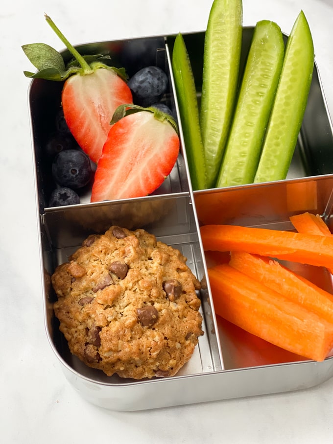 chunky chocolate chip oat cookies in a metal lunch box with strawberries, blueberries, sliced cucumber and carrot batons.