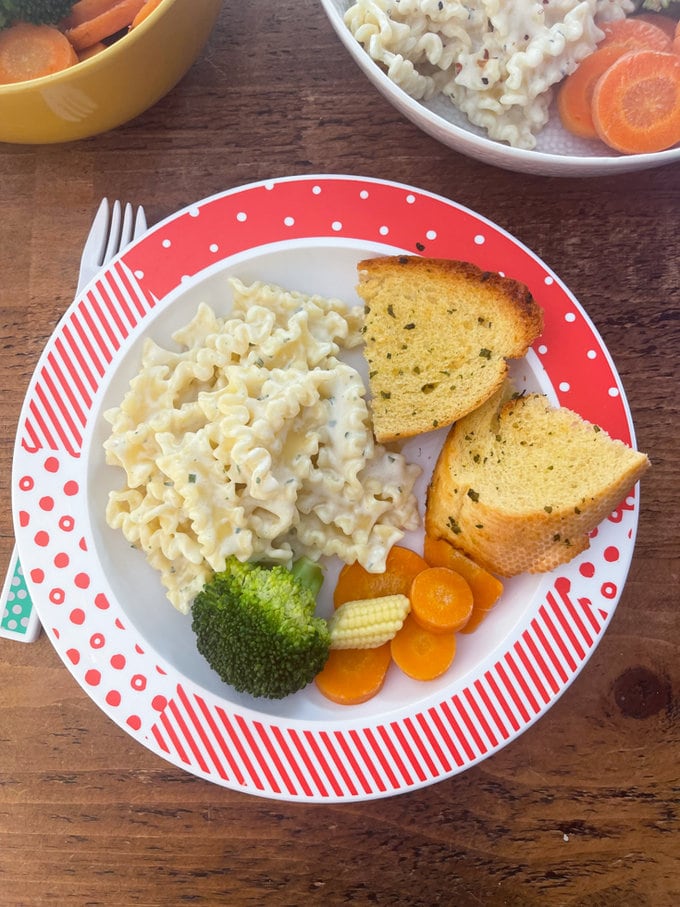 Boursin pasta served for kids with garlic bread and some vegetables on a red and white patterned plate