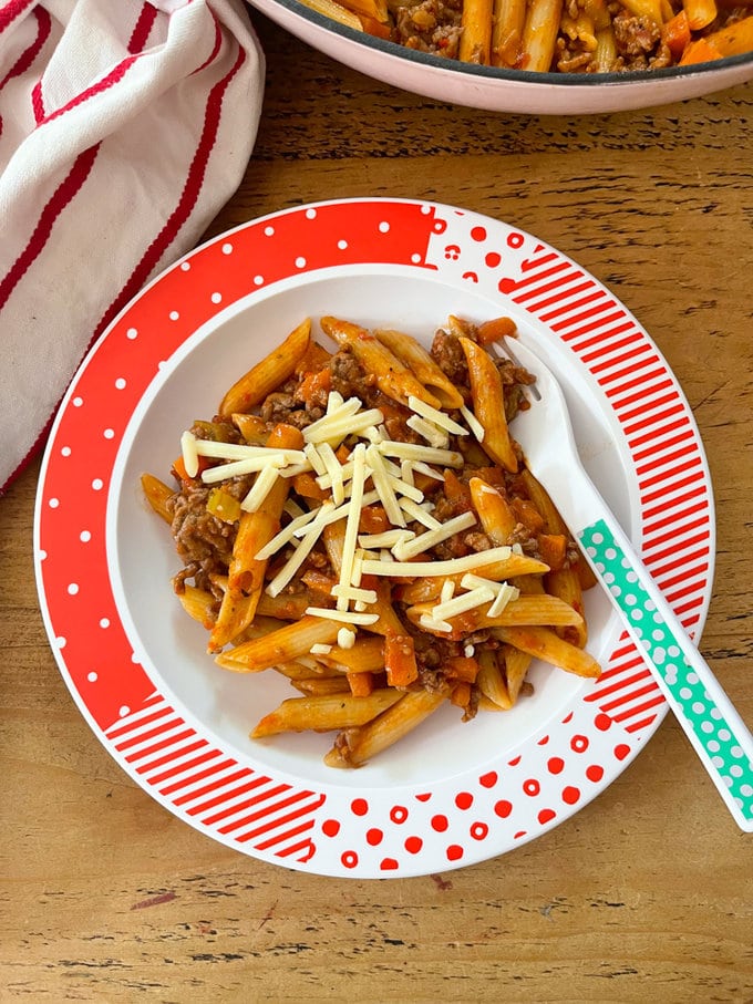 a kids serving of the meal in a red and white patterned bowl garnished with grated cheddar cheese