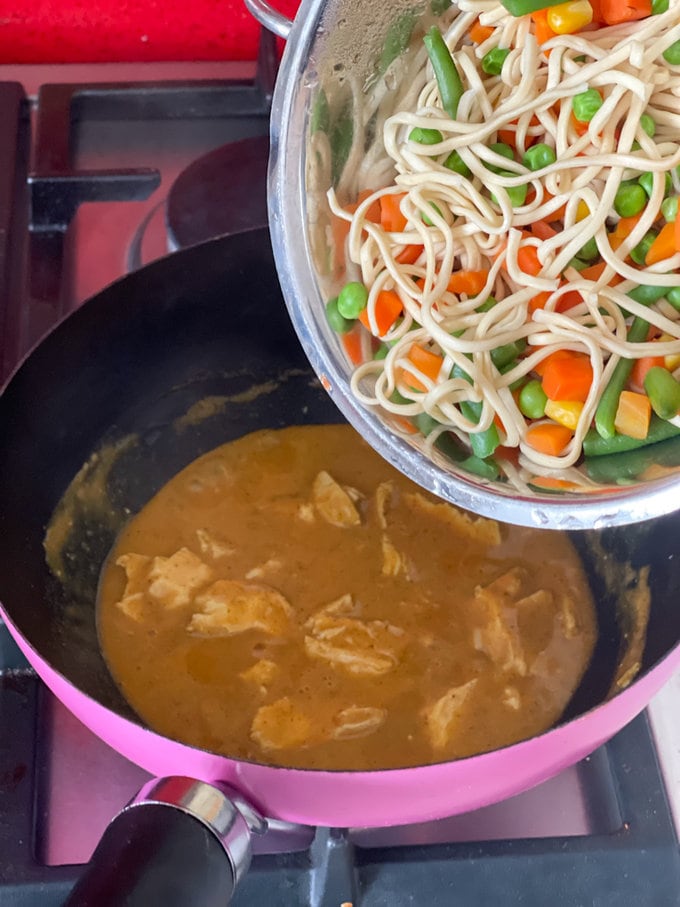cooked noodles and vegetables being added to the pan of chicken and curry sauce.