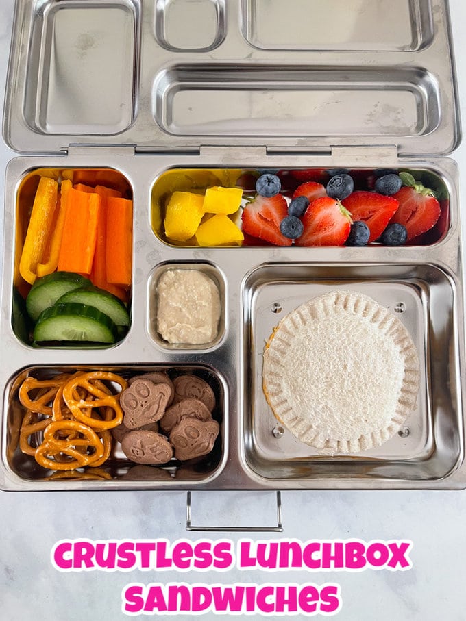 A lunch box with stainless steel compartments containing a crustless sandwich, chopped fresh fruit and vegetables, and some pretzels and mini chocolate chip cookies