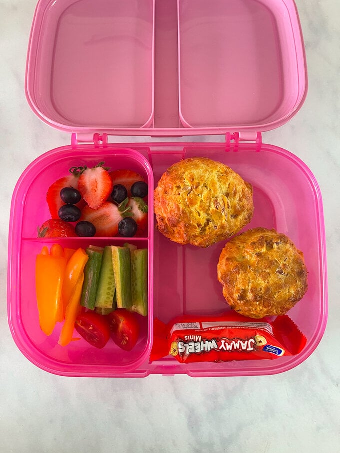 Ham and cheese muffins presented in a pink lunchbox alongside fruit and vegetable snacks.