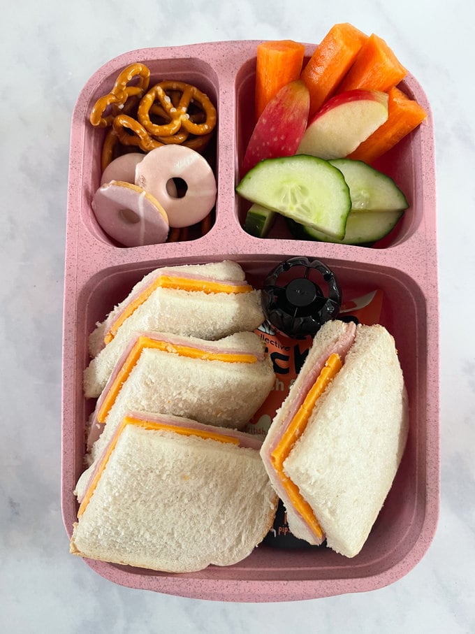 Cheese and ham sandwiches cut into four squares and presented in a pink lunchbox along with other snacks.
