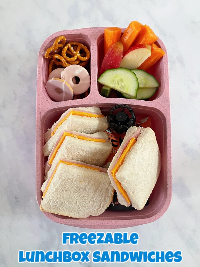 How to freeze sandwiches, defrosted and presented in a light pink three section lunchbox along side fresh chopped fruit and veggies, with a sweet treat iced biscuit.