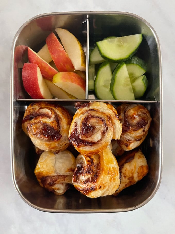 six mini jam pinwheels presented in a stainless-steel lunchbox along with chopped apple and cucumber.