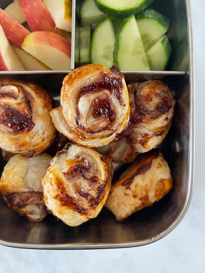 Mini Jam Pinwheels in a compartment lunchbox with slices of chopped red apple and cucumber.