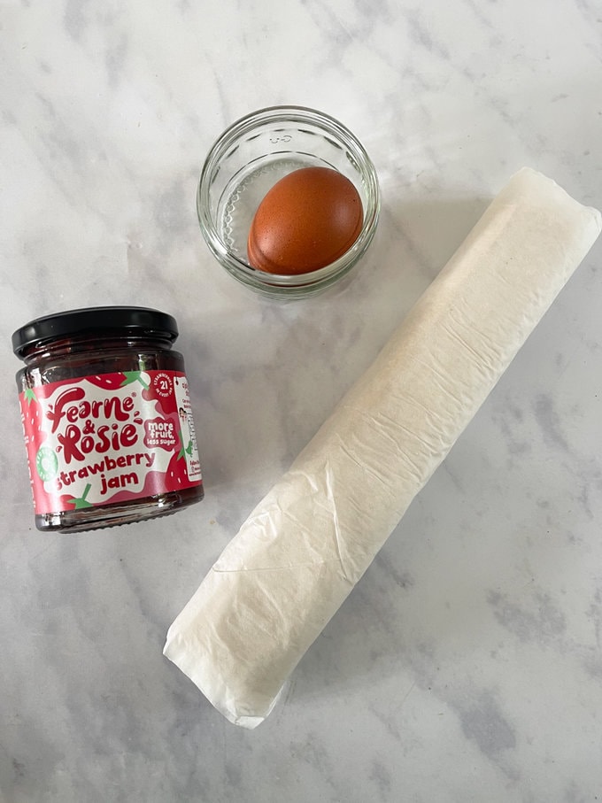 Strawberry jam, pastry and egg laid out on worktop.