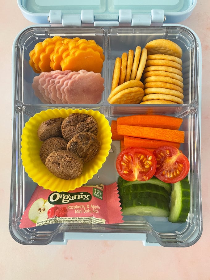 Lunchables presented in a sectioned lunchbox, in one section there is shaped ham and cheese slices, cheese crackers in another, and the last section filled with chopped and sliced fresh veggies, and mini chocolate chip cookies.