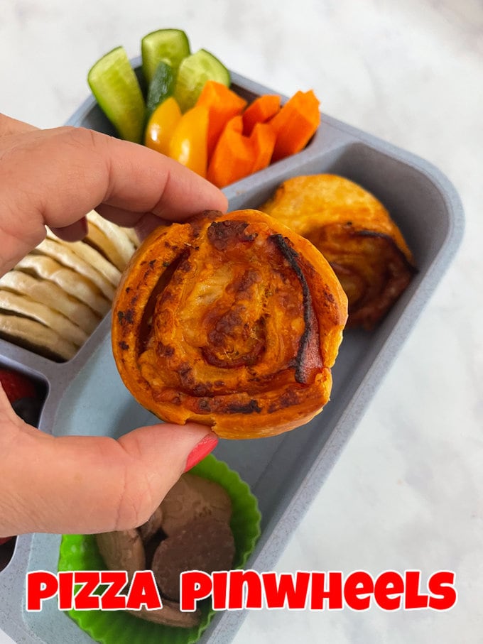Two pizza pinwheels presented in a sectioned lunchbox,served with fresh chopped veggies and rice crackers.