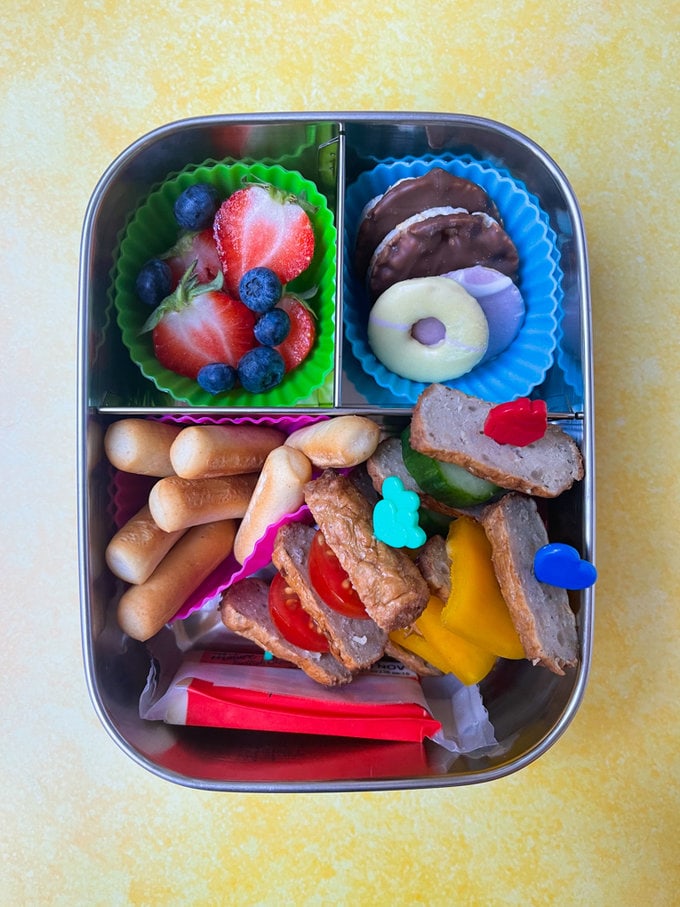 Sausage and vegetable skewers presented in a metal style lunchbox along with breadsticks, cheese, and sweet snacks.