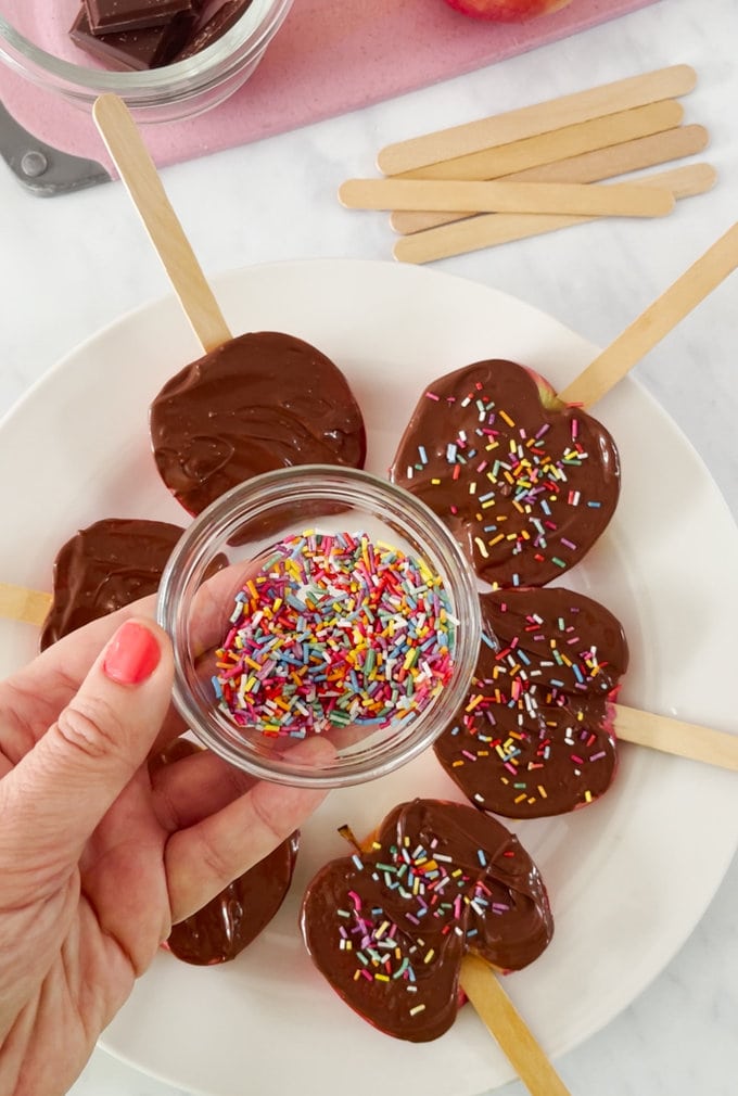 Chocolate apple slices on a white plate and colorful sprinkles being added. 