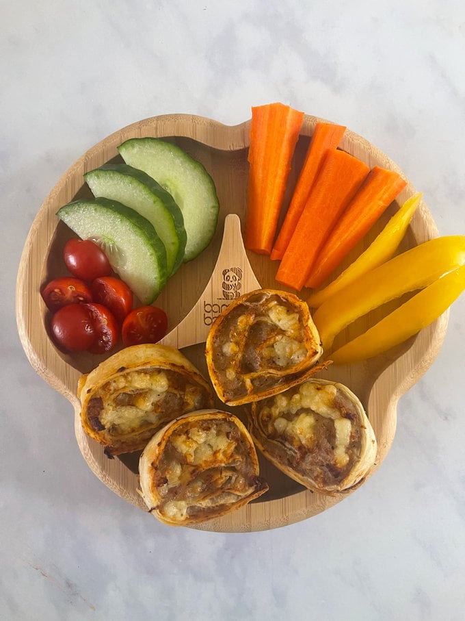 Golden brown cheese and sausage pinwheels presented on a child's bamboo plate, presented with fresh colorful cherry tomatoes, cucumber, carrot sticks and yellow peppers.