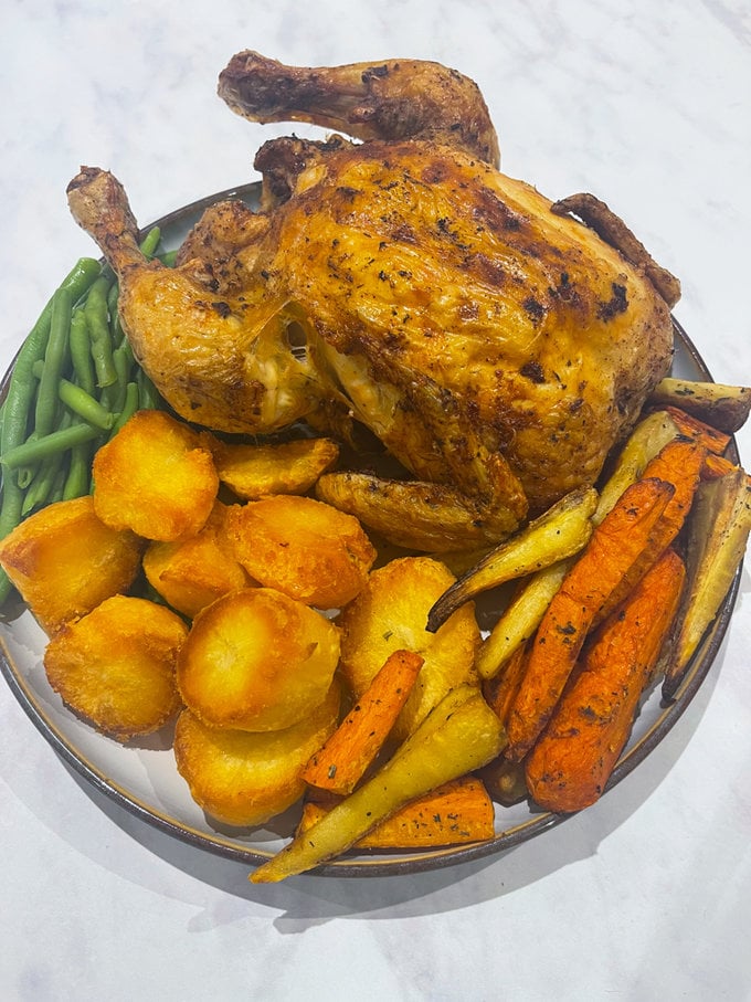 Airfyer whole chicken presented on a large plate and garnished with crispy roast potatoes and a selection of vegetables.