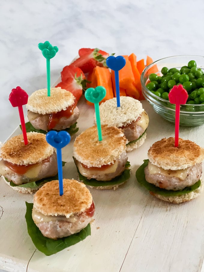 Five mini chicken burgers presented on a large plate, garnished with lettuce, melted cheese and tomato ketchup, and held together with bright pick sticks. served alongside peas, raw carrots and strawberries.