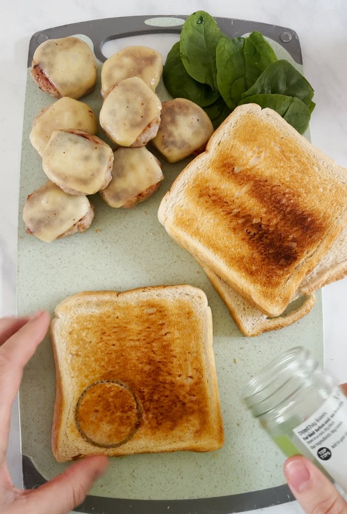 Cooked mini burgers with melted cheese on top, alongside lettuce and three pieces of toast. ready to be constructed on a chopping board.