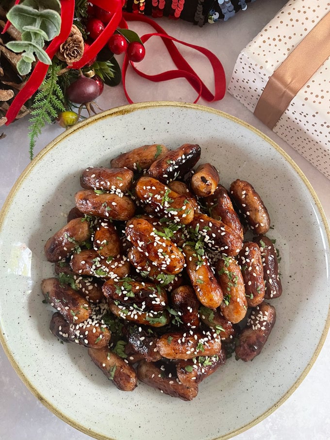 sticky asian cocktail sausages served in a ceramic bowl and topped with sesame seeds and chopped coriander