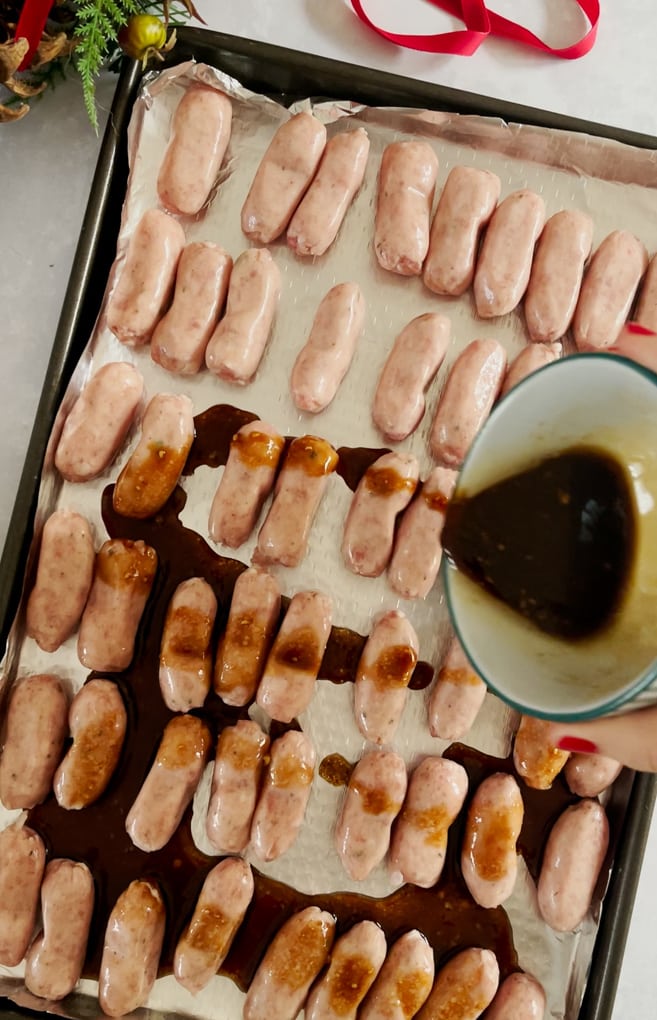 the glaze being poured over the raw cocktail sausages on the baking tray.