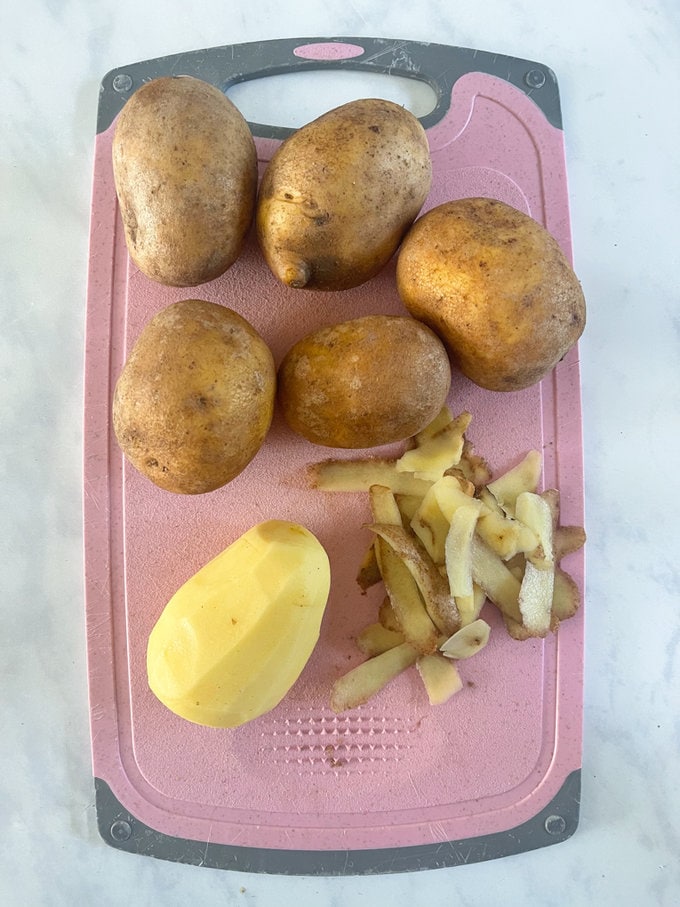 Five potatoes on a pink and grey plastic chopping board ready to be sliced.
