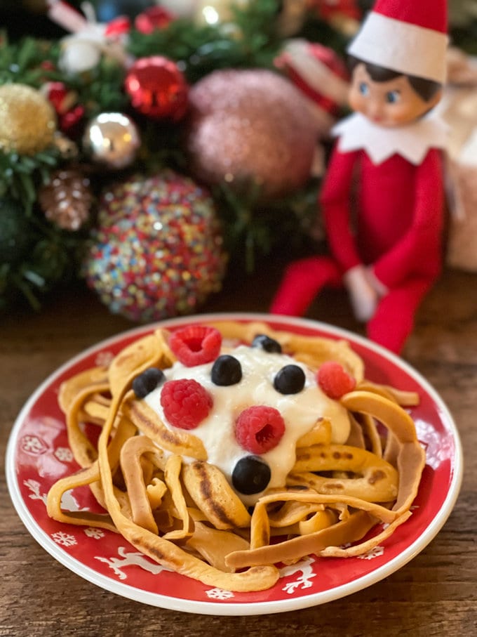 Buddy the elf sitting next to a Christmas themed red plate, full of Elf spaghetti. Garnished with Greek yogurt and berries.   