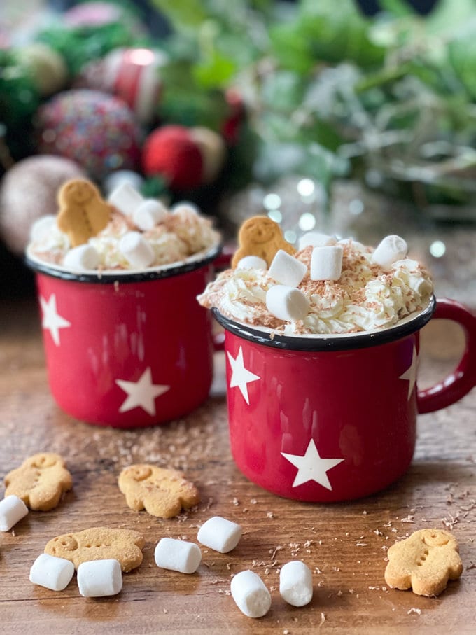 Gingerbread hot chocolate served in red cups with white stars. Garnished with whipped cream, grated chocolate flakes, mini marshmallows and gingerbread men.