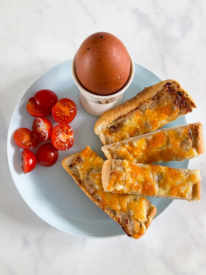 Airfryer Sausage and cheese soldiers arranged on a white round plate. With a cooked egg in  a egg cup. And cherry tomatoes cut into halves.