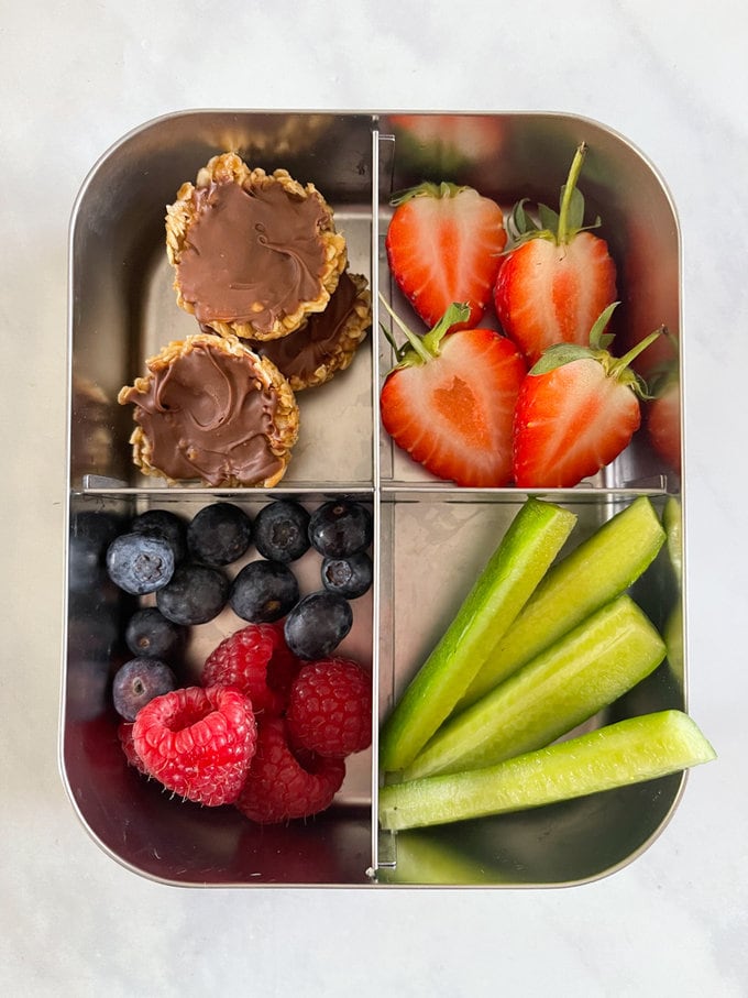 Butter oat cakes arranged in a metal lunchbox with four compartments, accompanied with fresh berries and cucumber sticks. 