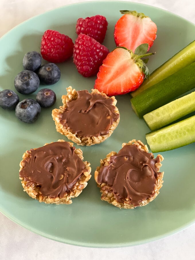 Three Butter oat cups displayed on a plate along with fresh berries and cucumber sticks.