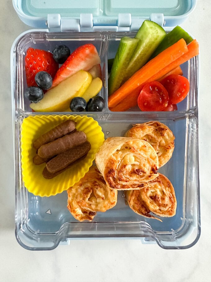 Four Airfryer Chicken & Cheese Pinwheels served in a child's divided lunchbox along side fresh,chopped fruit and veggies, with some mini chocolate fingers.