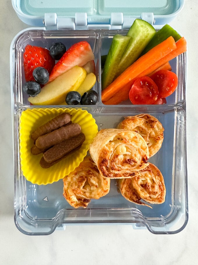 Airfryer Chicken & Cheese Pinwheels in a compartment lunchbox with fresh fruit, chopped veggies and mini chocolate fingers