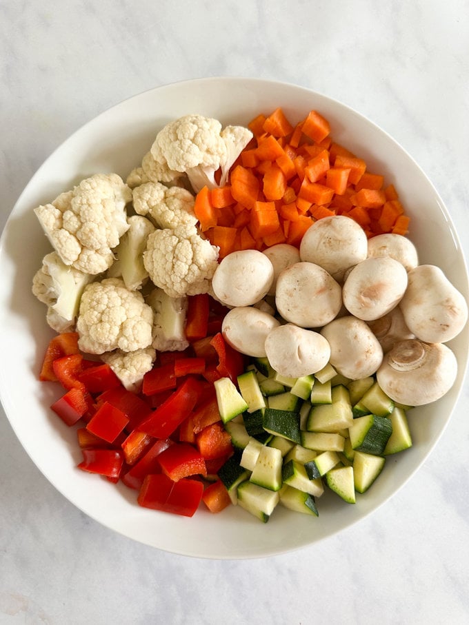 A white round bowl full of chopped Carrots,courgette,Cauliflower,pepper and mushrooms.