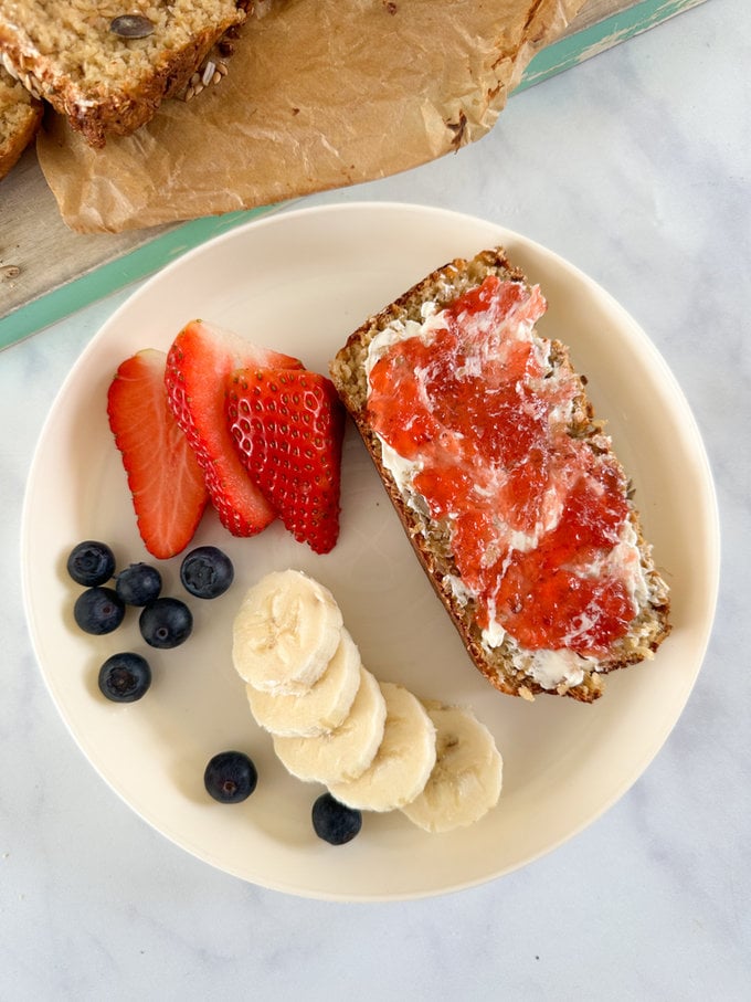 a slice of porridge bread topped with butter and jam in a white bowl served with sliced banana, slice strawberries and fresh blueberries