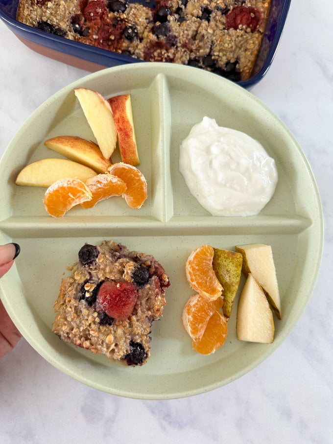 A small portion of Baked berry oat bar presented on a round plastic divided plate along with fresh fruit and yogurt. 
