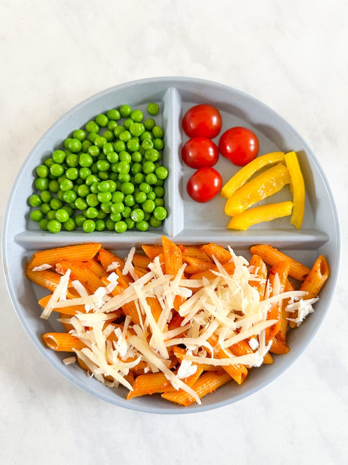 Child's round divided plate, with pasta,peas,cherry tomatoes and sliced peppers. Pasta garnished with grated cheddar and feta cheese  