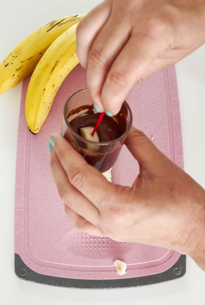 Banana slice being dipped into a small pot of melted mil chocolate.