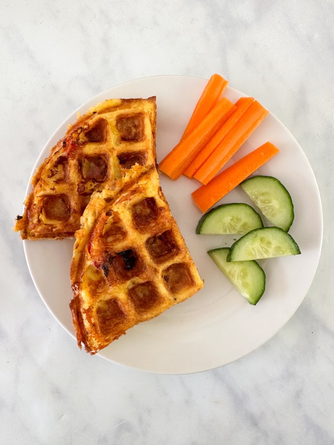 Waffle omelette presented on a round white plate, and served with carrot sticks and sliced cucumber.