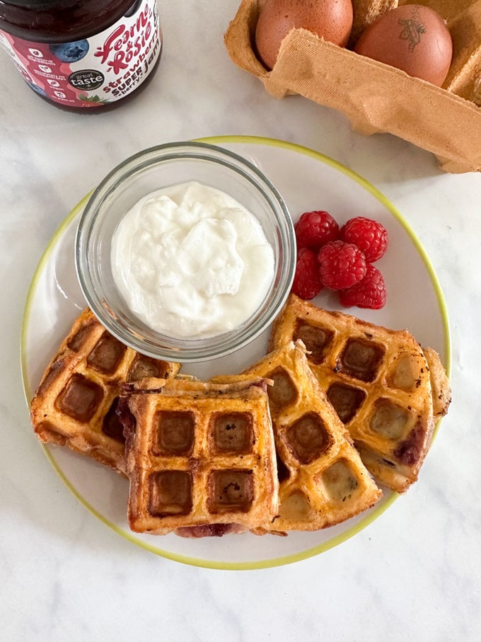 Waffle french toast presented on a small round plate with fresh raspberries and a small side bowl of Greek yogurt. 