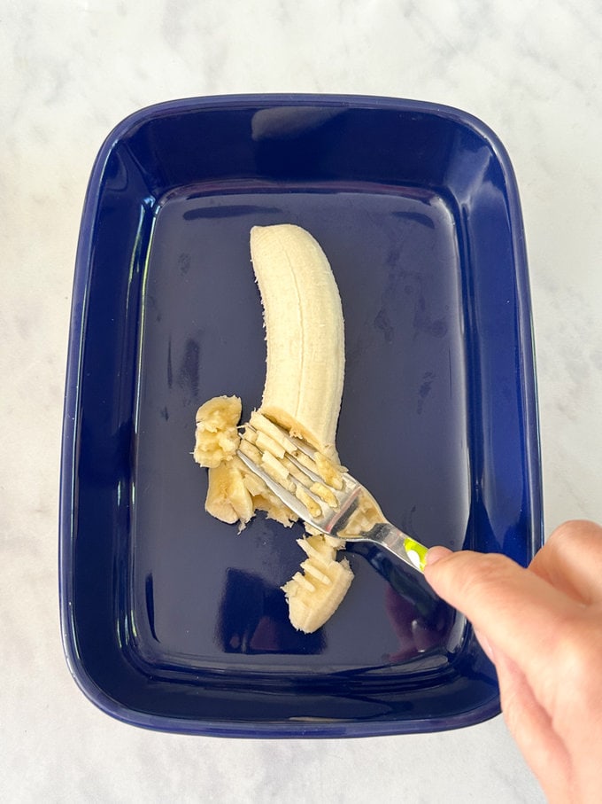 Whole Banana being mashed up with a fork in a blue dish.