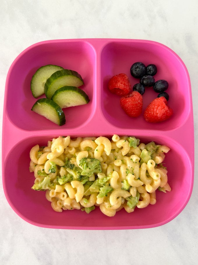 One pot Broccoli mac and cheese presented on a bright pink,divided child's plate, served with fresh berries and cucumber slices.