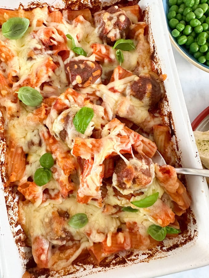 Easy meatball pasta bake served in a large white oven dish, finished with melted cheese and garnished with basil leaves. 