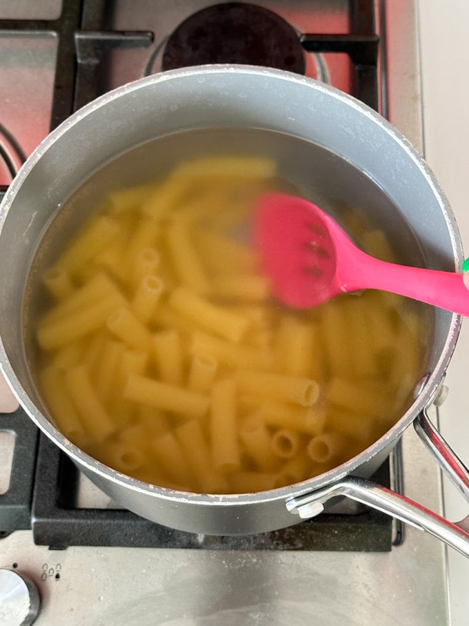 Pasta tube in a large saucepan, being stirred with a bright pink spoon.