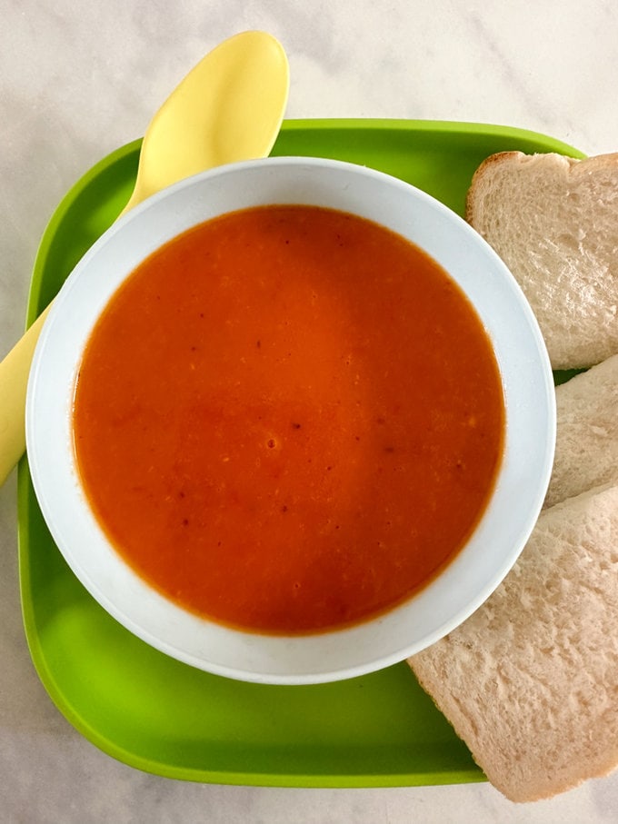 Roasted tomato & red pepper soup served in a small white bowl, accompanied by a slice of white bread. 
