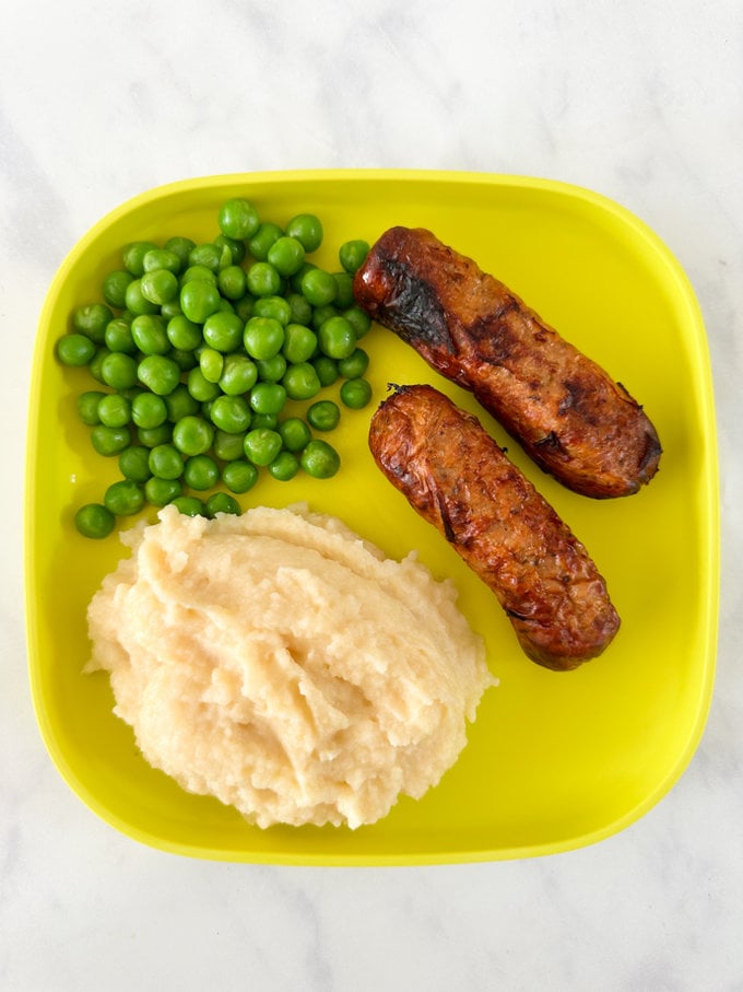 5 minute tinned mashed potato served alongside two sausages and green peas on a bright yellow plastic plate.