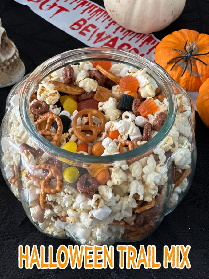 Fun Halloween Food Ideas For Kids -Halloween Trail Mix in a large glass pumpkin shaped jar with Halloween decorations in the background.
